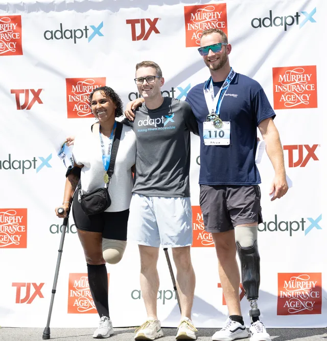 Three athletes wearing race medals at a sponsor backdrop, one on a forearm crutch and one with a prosthetic leg