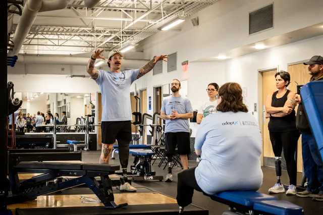 A coach with a prosthetic leg, arms raised, leading a clinic session in a gym as other athletes on prostheses watch