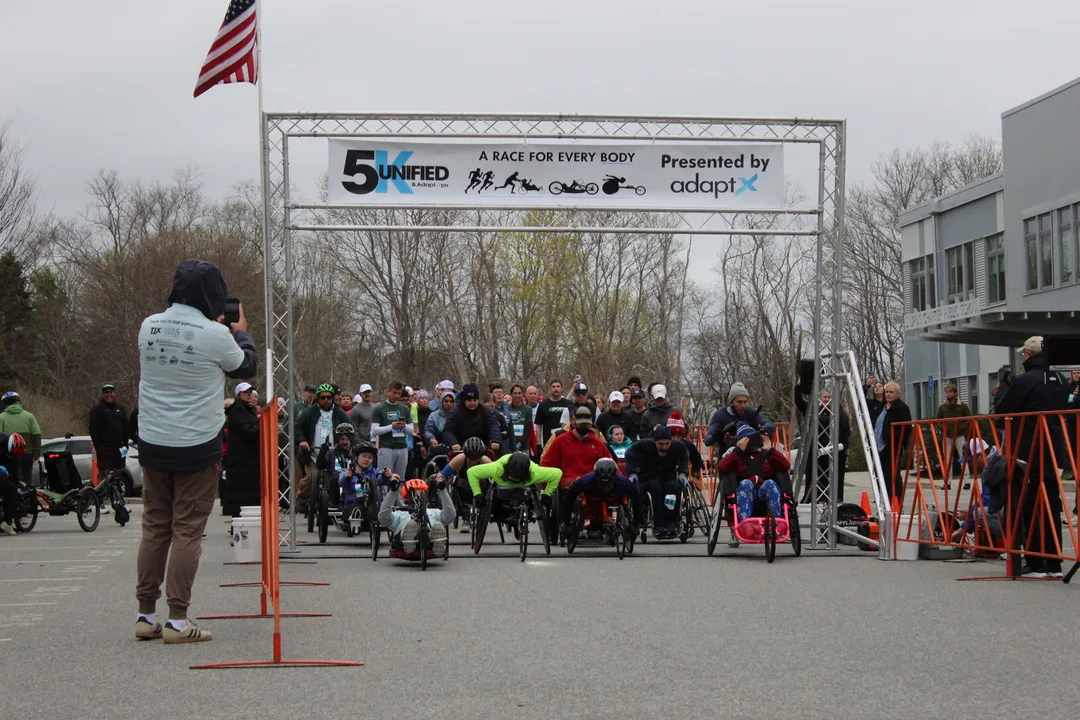 A crowd of runners and wheelchair racers packed under the Unified 5K start arch on a tree-lined street