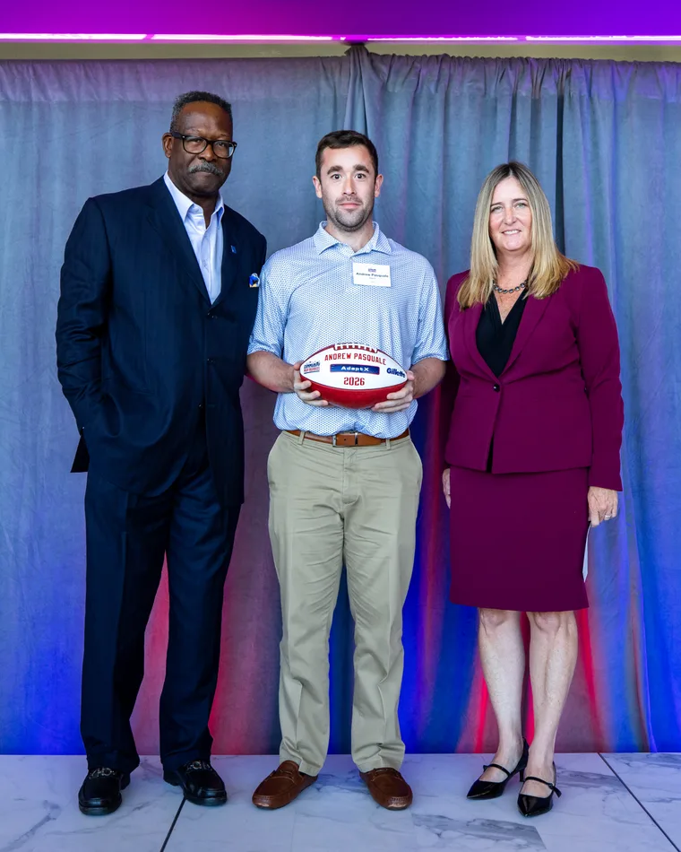 A man in a light blue polo stands next to two Patriots representatives while holding a football that says Andrew Pasquale and AdaptX.